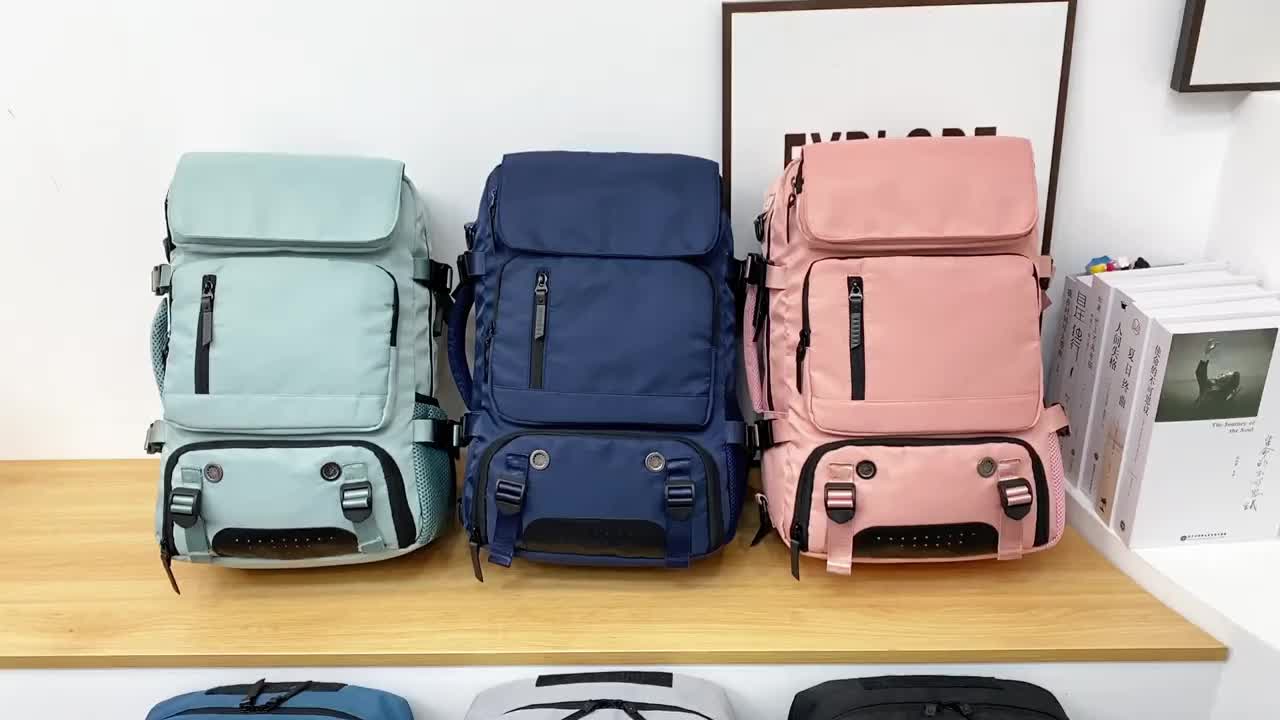 Three backpacks in light blue, navy, and pink on a wooden surface with a white wall background.